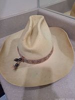 Front and angled views of vintage felt cowboy hat showing the 4" brim, pinched crown, and ornate patterned fabric hatband with bead and feather detail. Visible dirt and wear marks on felt surface.