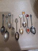 Nine vintage spoons and one fork arranged side by side on a beige surface, showing various handle designs and sizes.