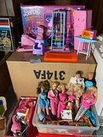 Overview of Barbie dolls, Ken, Skipper, playsets including gym and news themed boxes, and small accessories in a plastic bin and arranged on a box.