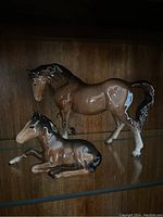 Pair of Beswick horse figurines on a glass shelf, mother horse standing and baby horse lying down.