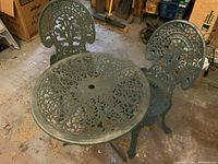 Two matching green aluminum bistro chairs positioned around round table with cutout pattern and umbrella hole.