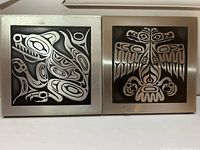 Two silver-tone West Coast Indian art plaques with wolf and eagle designs on black background, framed with silver border.
