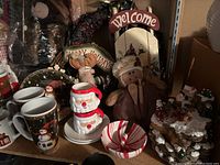 Shelf with ceramic holiday mugs featuring snowman designs, Santa Claus-themed ceramic bowl, peppermint swirl ceramic bowl, holiday fabric figurines including a gingerbread person and reindeer, and snowy tree candles with red and white accents.