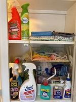 Photo of upper and middle cabinet shelves showing various cleaning sprays, bottles, and folded cleaning cloths and towels.