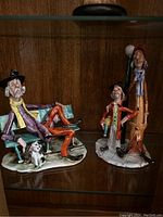 Two clown figurines displayed on glass shelf with wood background.