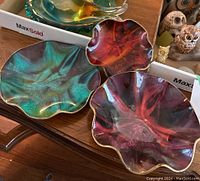Three wavy-shaped colored glass decorative bowls in teal, red, and purple with gold edging on a wooden table.