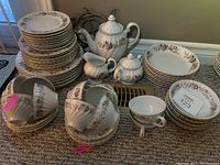 Full view showing stack of plates, bowls, cups, teapot, milk jug, sugar bowl, and metal trivet, arranged on carpeted floor