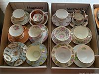 Two lot trays each holding six cups and six saucers with floral patterns and gold accents.