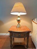 Full view showing medium wood-tone end table with carved details and brass-tone table lamp with cream shade on top, plugged in and lit under neutral wall and hardwood floor