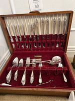 Wooden box open showing silver plate flatware items neatly stored in red velvet slots with cream silk backing. Multiple butter knives, forks, spoons, and a serving spoon visible.