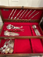 Wooden flatware box with red velvet lining containing multiple pieces of silverplate flatware including knives, forks, and spoons neatly arranged.