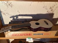 Maccaferri Islander Deluxe ukulele lying on its original box on a wooden shelf with floral wallpaper background