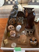 Photo of multiple assorted owl figurines arranged in a cardboard box on a wooden table, showing wood carved, glass, and painted resin materials.