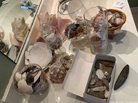 Overhead view of glass apothecary jar, cylinder vase, compote dish, wicker basket filled with shells, large conch shell, and box of arrowheads