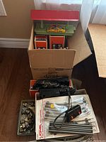 Lionel 027 gauge electric train items including boxed tracks, locomotive, and train station inside a cardboard box.