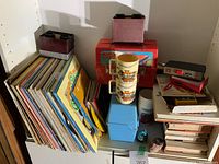 Collection of vinyl records and albums leaning vertically, with stacked vintage mugs, View Master toys, lunchboxes, and various small items.