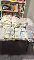 Photo showing several folded bedding items tied with ribbons arranged on the floor in front of bookshelf.