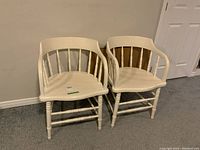 Two white painted wood Windsor style chairs side by side against a wall on carpeted floor.