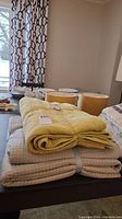Stack of three throw blankets on a table with lamps and dishes in background. The top blanket is apple green/yellow and folded with a label visible.