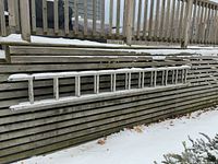 Image shows full length of aluminum extension ladder resting horizontally against a wooden slatted wall, outdoors with snow on the ground and on parts of the ladder.