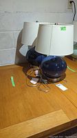 Two matching small table lamps with blue round bases and white shades on wooden table against cinderblock wall.
