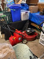 Frigidaire red push lawn mower with two red plastic gas cans beside it in a storage area with cluttered background items.