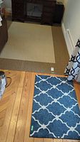 Shows the large brown area rug with the small blue door mat in the foreground on a wood floor.