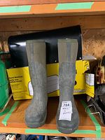 Two tall Muckmaster boots standing in front of a black aluminum rural mailbox box packaging on a shelf.