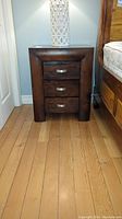 Front view of one nightstand showing three drawers with silver half-moon handles, visibly dark wood finish, sitting on wood floor.