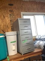 Gray metal rolling cabinet with six drawers next to white and turquoise waste bins on wooden shelf