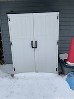 Front view of white resin shed with double doors and black handles on snow-covered ground