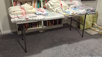 White folding table with assorted fabric bundles on top; carpeted floor and bookshelf in background.