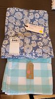 Photo showing a bundle of blue floral patterned 100% cotton table runners with a tag noting 6 blue and 5 green runners, measurements 12x80 and 11x82.