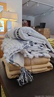 Stack of four throw blankets: two cream and navy tasselled throws on top, two pale mustard cotton throws on bottom.