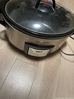 Photo of an oval-shaped stainless steel Crock-Pot digital slow cooker with a glass lid and black handle. Digital control panel and power cord visible.