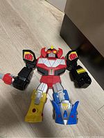 Photo of a large Power Rangers Megazord toy figure lying on floor, showing front view with colorful dinosaur-themed legs and red and black torso and arms.