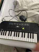 Casio SA-46 mini keyboard on a white bed showing front with 32 small keys, control buttons, numeric keypad, LCD screen, and attached power adapter cable.