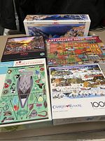 Five 1000-piece puzzles displayed on table with visible box art of each puzzle. Coastal panoramic scene, street scene, nature and wildlife illustration, and winter town scenes are visible.