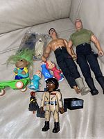 Single photo showing a variety of toys including two muscular male action figures, a Ghostbusters figure, three Alvin and the Chipmunks figures, a Troll doll in packaging, and a green skateboard.