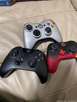 Three Xbox controllers on a couch: a silver Xbox 360 Halo Reach controller with colored buttons, a black Xbox One controller, and a black-to-red gradient Xbox One controller with a red D-pad.