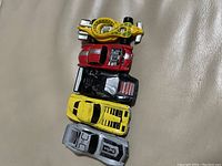 Five vintage toy cars arranged vertically on a beige leather surface showing different colors including yellow, silver, red, black, and yellow-green with visible wear and use.