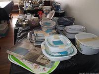 Photo showing multiple glass bowls, baking dishes, and platters arranged on tables, mostly white and clear glass, some with lids and floral patterns.