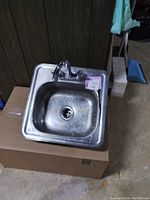 Square stainless steel bar sink with faucet resting on cardboard box, showing used condition with surface dirt and water spots.