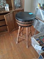 Bar stool showing black vinyl padded round seat, light wood legs and black metal accents beneath seat. Floor wood planks visible.
