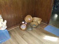 Five wicker baskets arranged on a wood floor against a wood-paneled wall under overhead light.