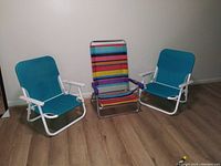Three low seating beach chairs arranged indoors on wooden floor: two blue fabric chairs with white metal frames and one multicolored striped chair with silver metal frame and purple armrest covers.