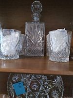 Lead crystal canister with stopper and four lead crystal beverage glasses on wooden shelf showing cut diamond and star patterns.