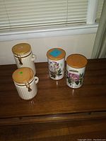 Four ceramic canisters on a wooden table against a window, two cream ridged ones with wooden lids and clasps, and two floral motif ones with wooden lids.