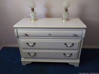White child's dresser with three drawers shown from front with two lamps on top