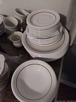 Stacked dinner plates, salad plates, bowls, cups, and saucers showing white ceramic with brown rim trim.